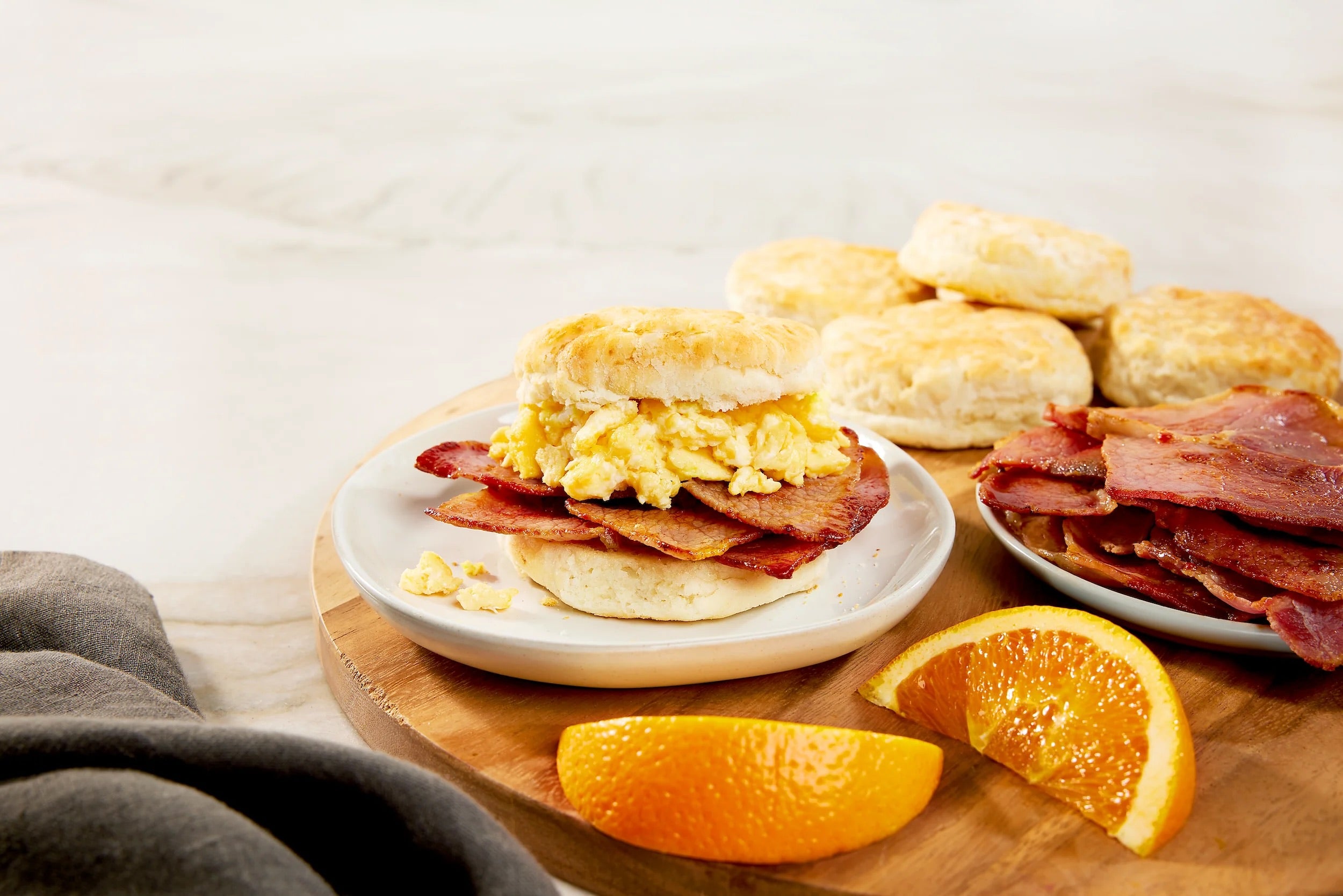 Breakfast setting with a biscuit sandwich, Suncrest Farms country ham, and orange slices on a wooden board.