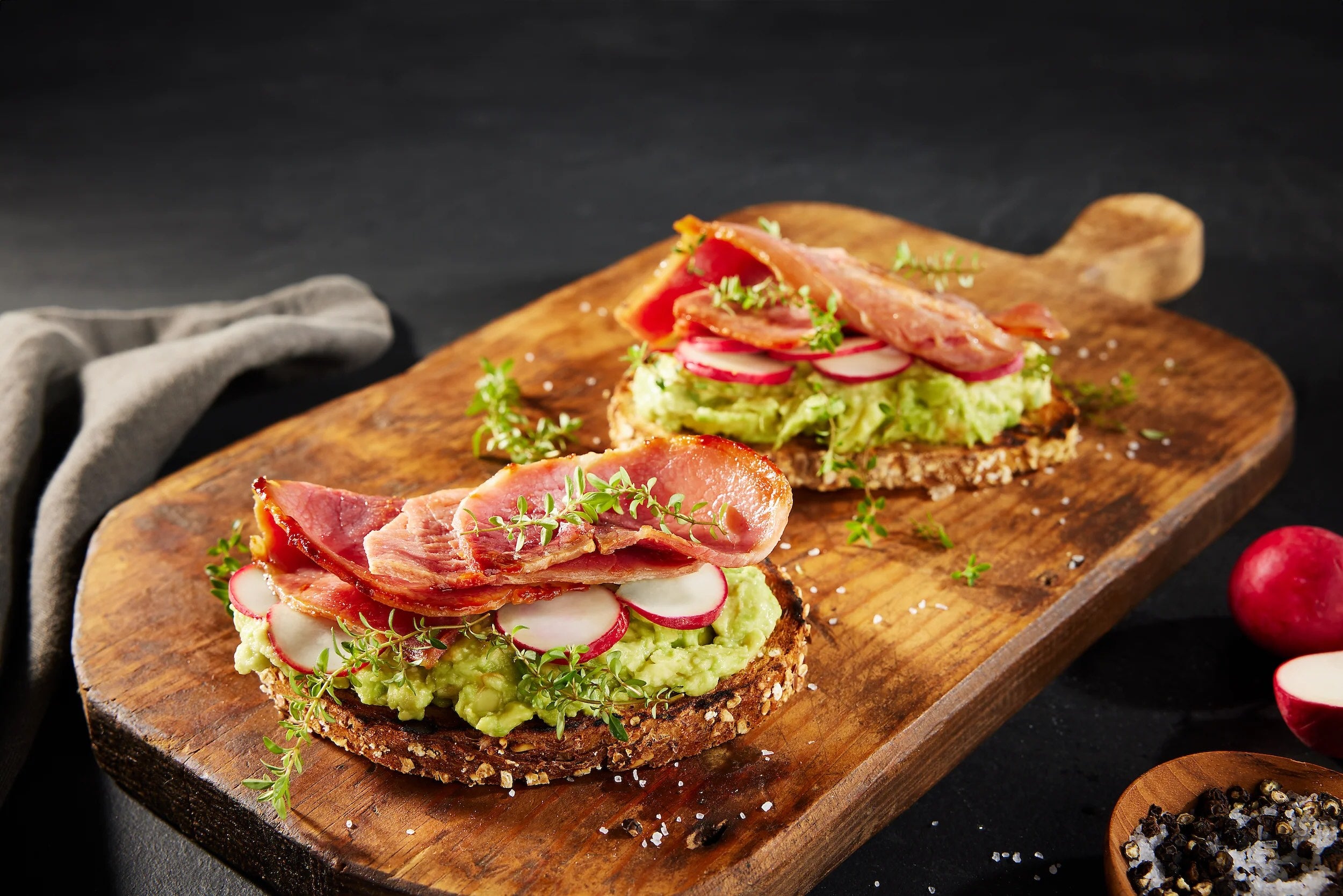 Two sandwiches with avocado, radishes, and Suncrest Farms country ham on a wooden cutting board.