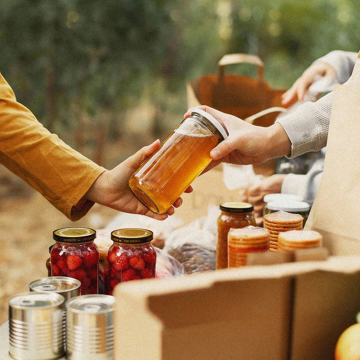 Person handing over a jar of honey to another person with a box of canned goods in the background.