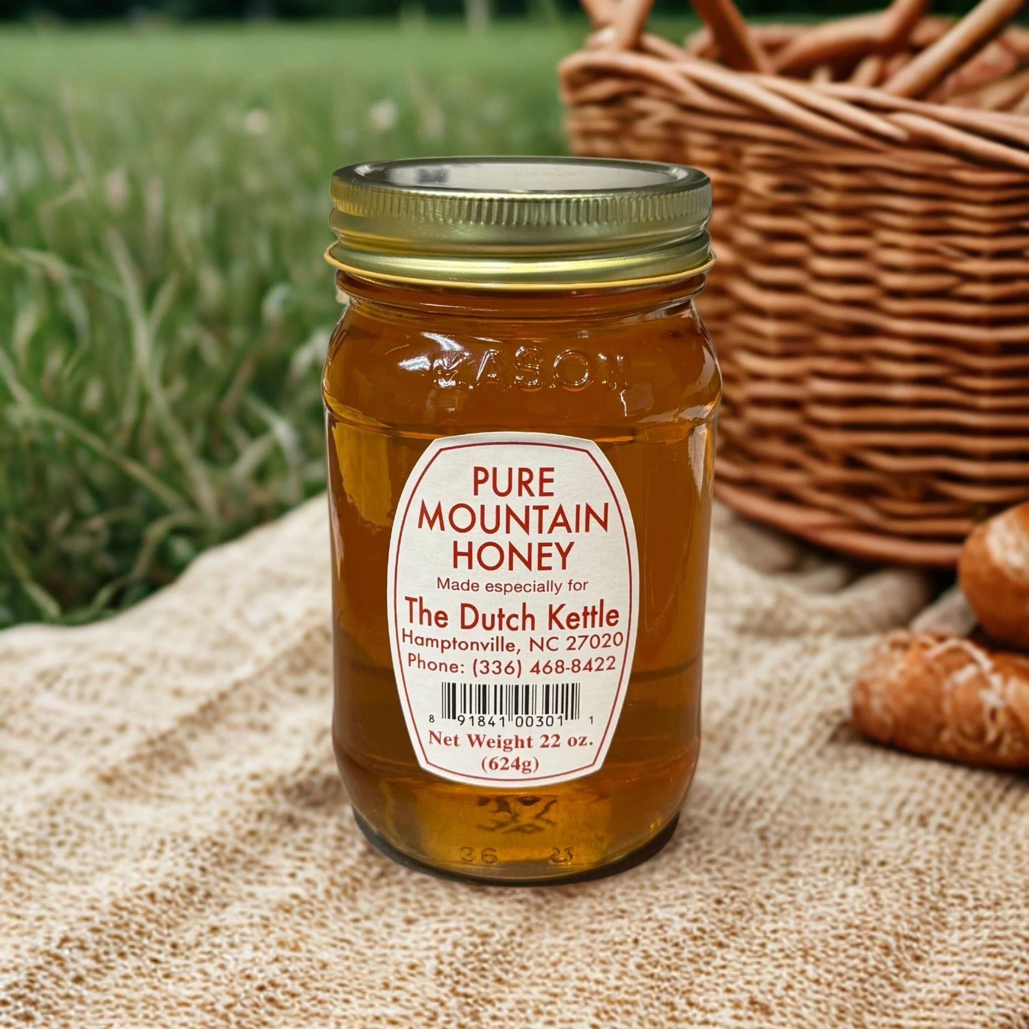 Jar of Dutch Kettle pure mountain honey with a label on a textured surface with a basket and bread in the background.