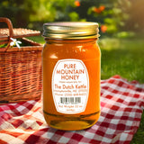 Jar of Dutch Kettle Amish Pure Mountain Honey on a checkered cloth with a picnic basket in the background.