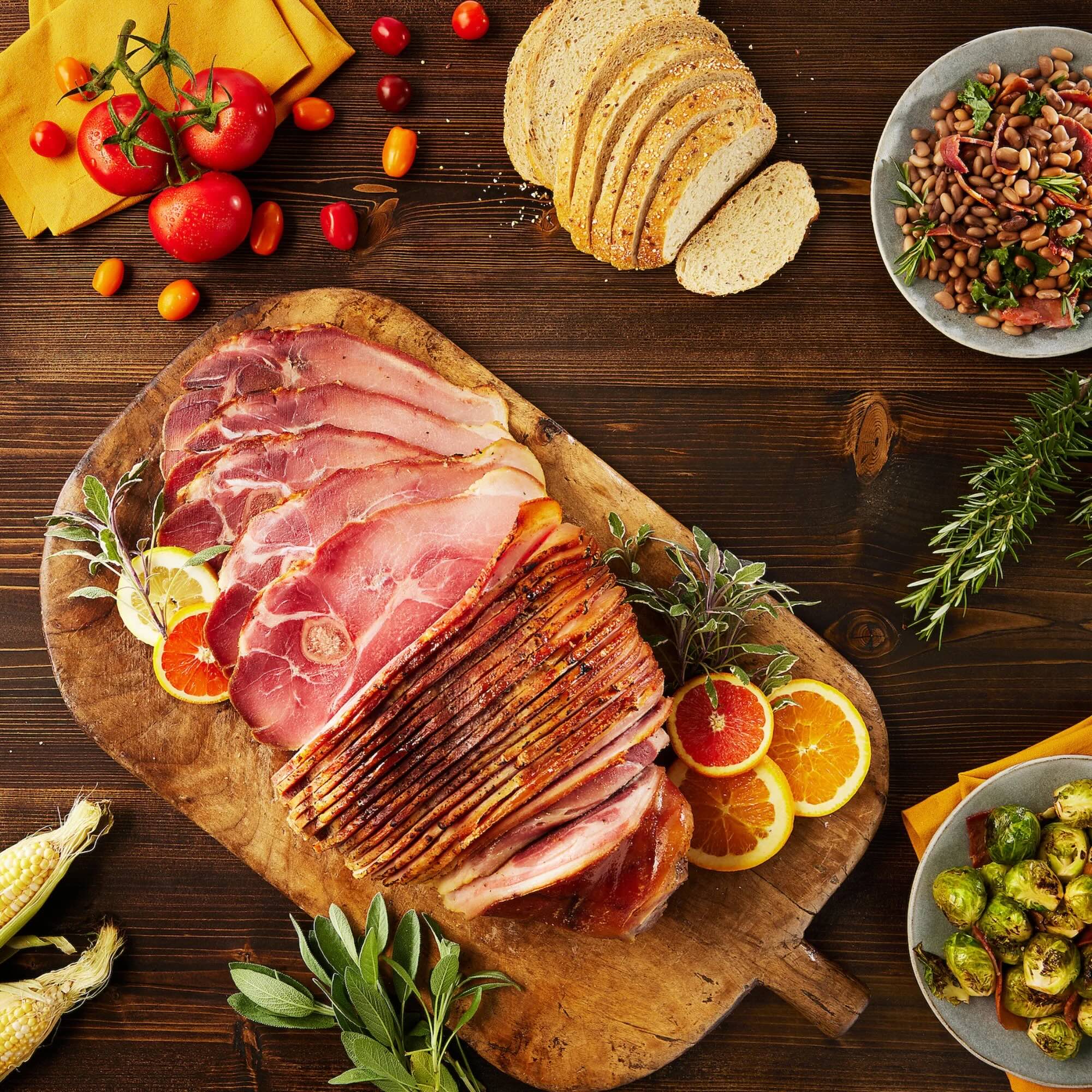 Wooden cutting board with Suncrest Farms sliced country ham, surrounded by vegetables and bread on a wooden table.