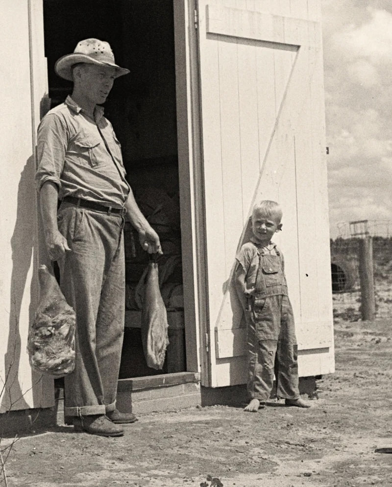 Black and white vintage photo of man and young boy leaning against barn door holding cured country hams in bags on a farm.