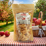 Perry Lowe Orchards Pink Lady dried appples in a tan package on a gingham picnic table with a picnic basket with apples in front of an apple orchard.