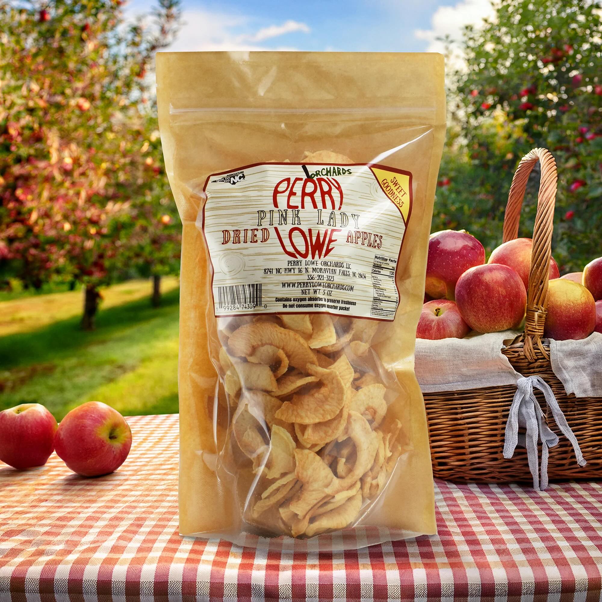 Perry Lowe Orchards Pink Lady dried appples in a tan package on a gingham picnic table with a picnic basket with apples in front of an apple orchard.