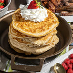 Stack of pancakes with whipped cream and a strawberry in a Stargazer 10.5 inch cast iron skillet, surrounded by berries and chocolate chips on a table.
