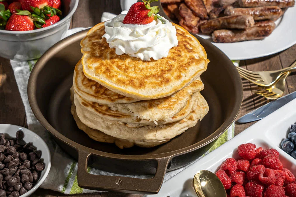 Stack of pancakes with whipped cream and a strawberry in a Stargazer 10.5 inch cast iron skillet, surrounded by berries and chocolate chips on a table.